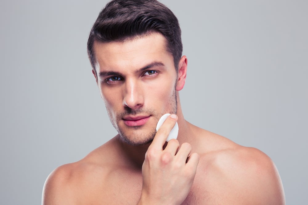 Man cleaning face skin with batting cotton pads over gray background